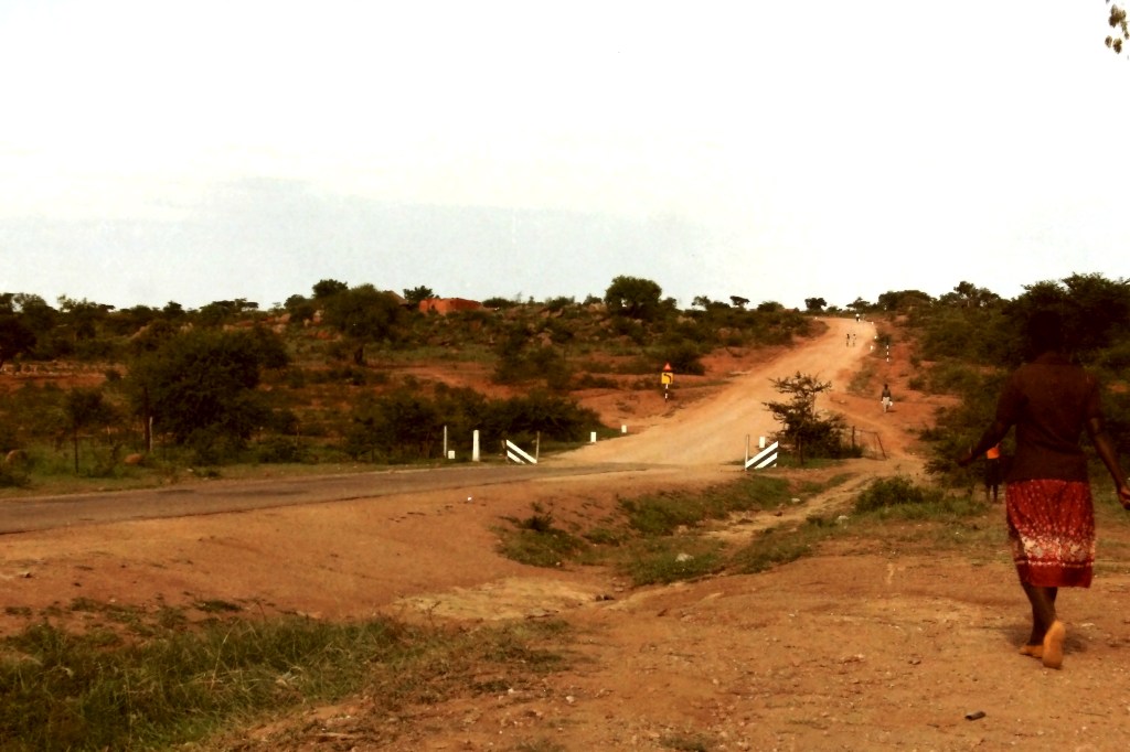 A stretch of Zimbabwe road, dirt roads, a cattle grid and road sign, with local residents walking.